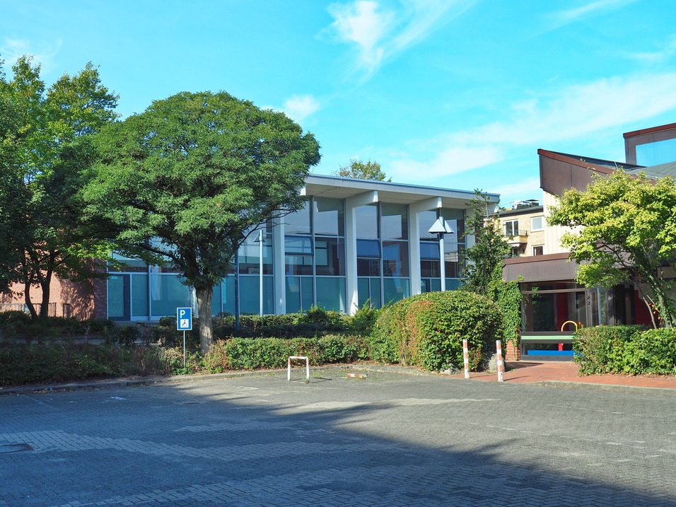 Moderne Geb&auml;ude mit gro&szlig;en Fenstern umgeben von B&auml;umen und einem gepflasterten Parkplatz. Blauer Himmel im Hintergrund.