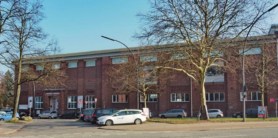 Ziegelbau mit Fenstern und B&auml;umen im Vordergrund, geparkt sind mehrere Autos an der Stra&szlig;e.