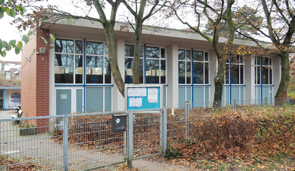 Ansicht eines modernen Geb&auml;udes mit gro&szlig;en Fenstern, umgeben von B&auml;umen und herbstlichem Laub, hinter einem Zaun.