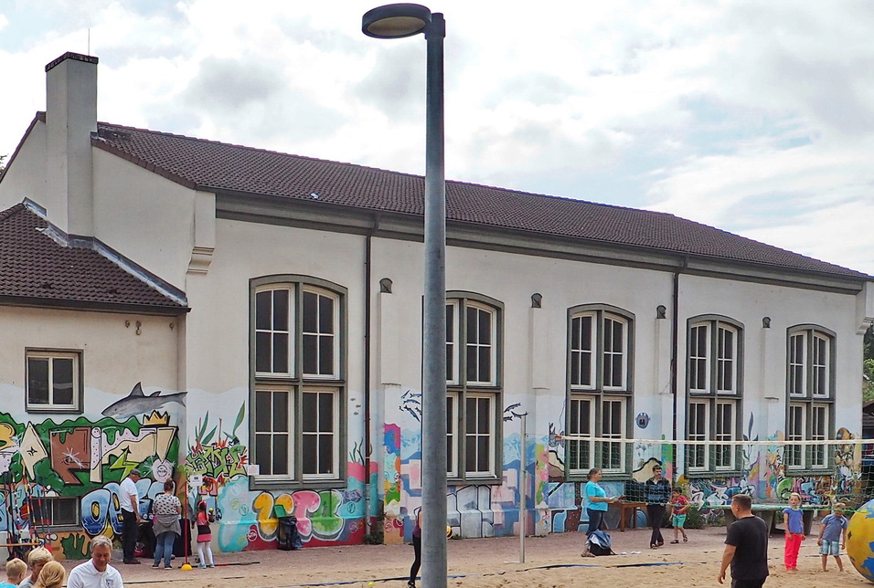 Graffiti-bemalte Wand einer Schule mit Kindern und Erwachsenen auf einem Spielplatz davor.