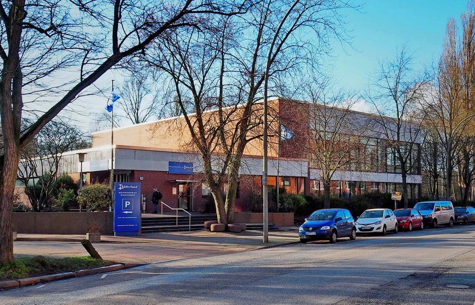 Eingangsbereich eines modernen, flachen Geb&auml;udes mit B&auml;umen und parkenden Autos auf einer ruhigen Stra&szlig;e.