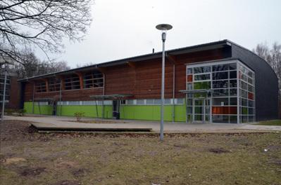 Moderner Sportgeb&auml;ude mit Holzverkleidung und gro&szlig;en Fenstern, umgeben von Gr&uuml;nfl&auml;chen und B&auml;umen.