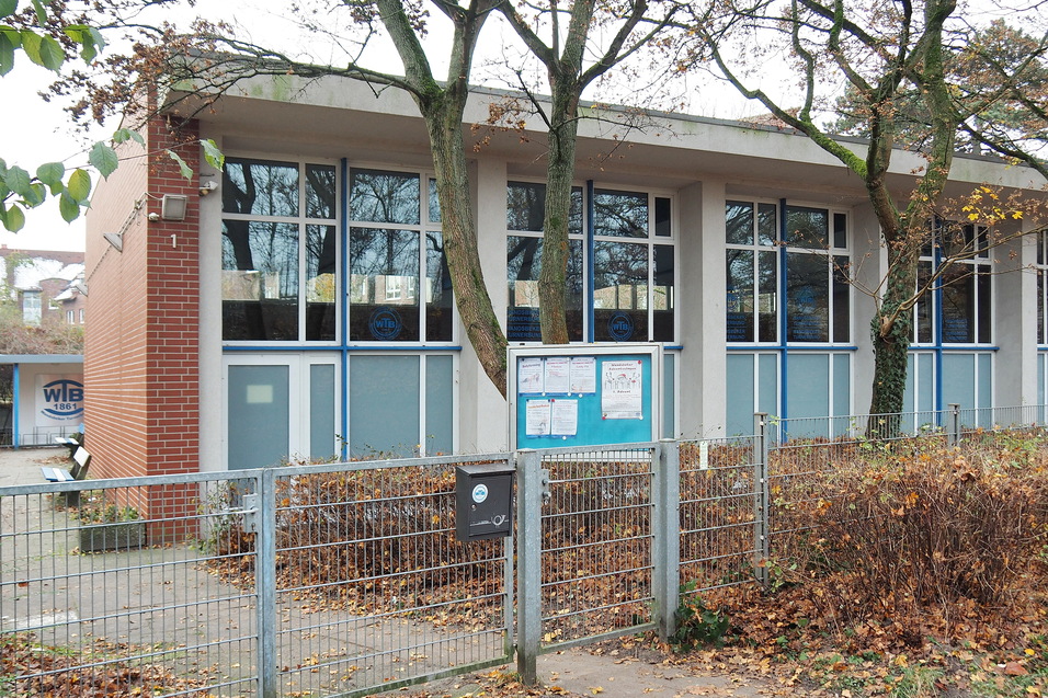 Ansicht eines modernen Geb&auml;udes mit gro&szlig;en Fenstern, umgeben von B&auml;umen und herbstlichem Laub, hinter einem Zaun.
