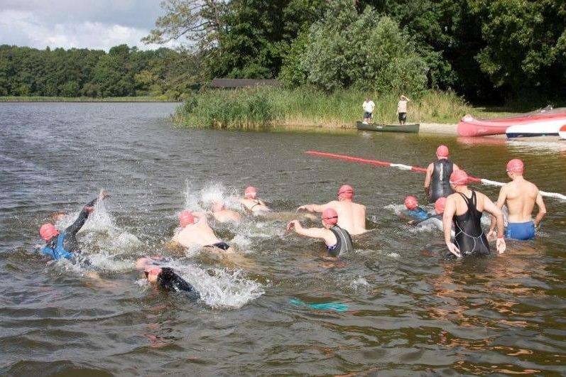 Gruppenschwimmen im See, Teilnehmer mit roten Schwimmhauben, im Hintergrund ein Boot und eine Wasserplane.