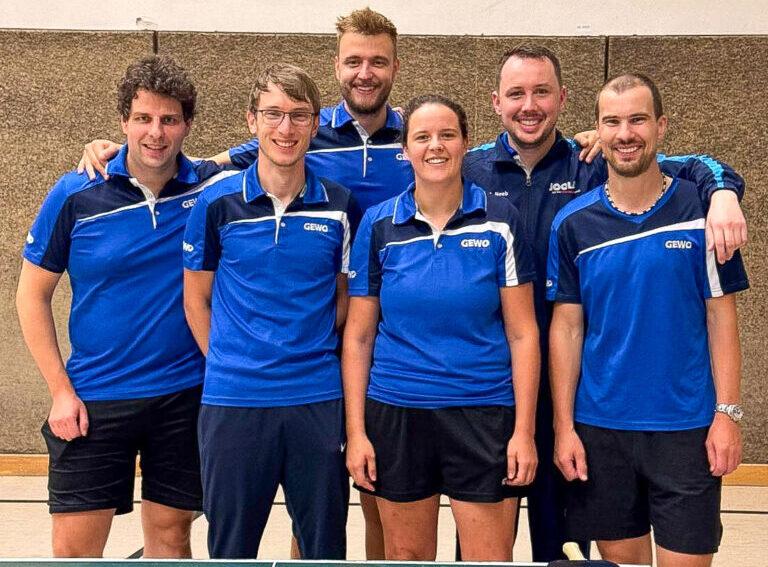 Sechs Tischtennisspieler in blauen Trikots posieren lächelnd vor einem Tischtennistisch.