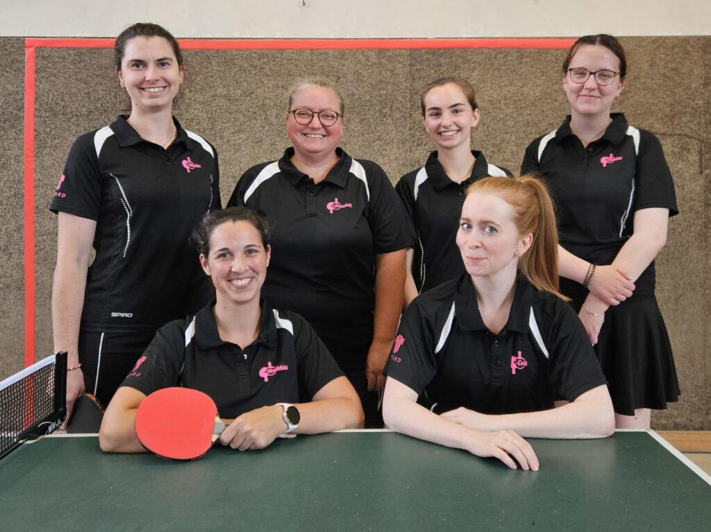 Gruppe von sechs Frauen in schwarzen Tischtenniskleidung, posierend vor einem Tischtennistisch mit Schläger und Ball.