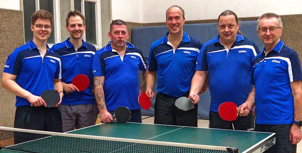 Sechs Männer in blauen Trikots stehen lächelnd vor einem Tischtennistisch mit Schlägern und Bällen.