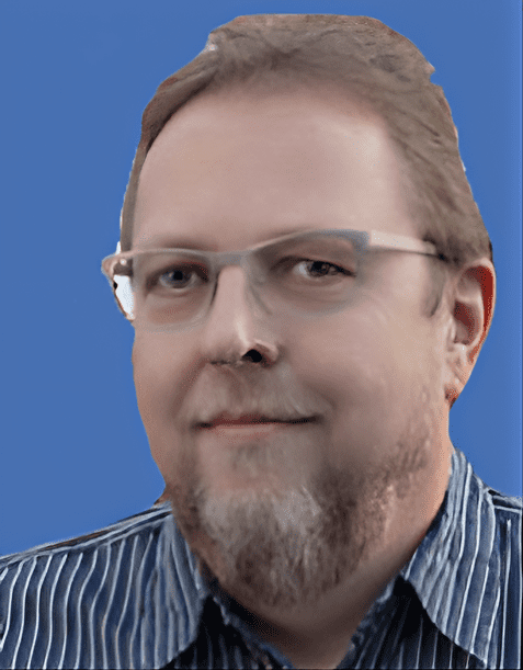 Ein Mann mit Brille und Bart lächelt vor einem blauen Hintergrund, getragenes gestreiftes Hemd.