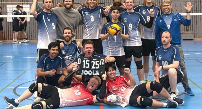 Gruppe von Volleyballspielern posiert jubelend in Sportkleidung, mit Ergebnisanzeige und Ball auf einem Hallenboden.