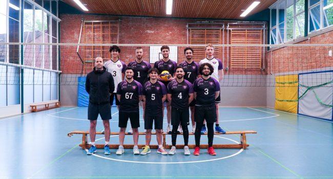 Männervolleyballteam posiert in Sportkleidung mit einem Volleyball in der Mitte auf einem Spielfeld in der Halle.