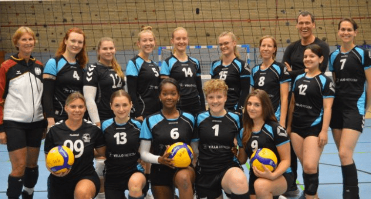 Gruppensicht auf ein Volleyballteam mit 15 Spielerinnen und Trainern in schwarzen und blauen Trikots mit Volleybällen.