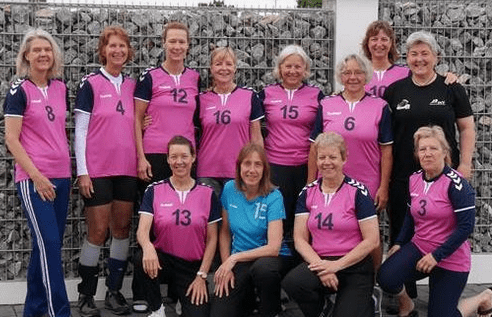 Gruppe von zwölf Frauen in pinken Sporttrikots, posierend vor einer Steinzäunwand, einige kniend, andere stehend.