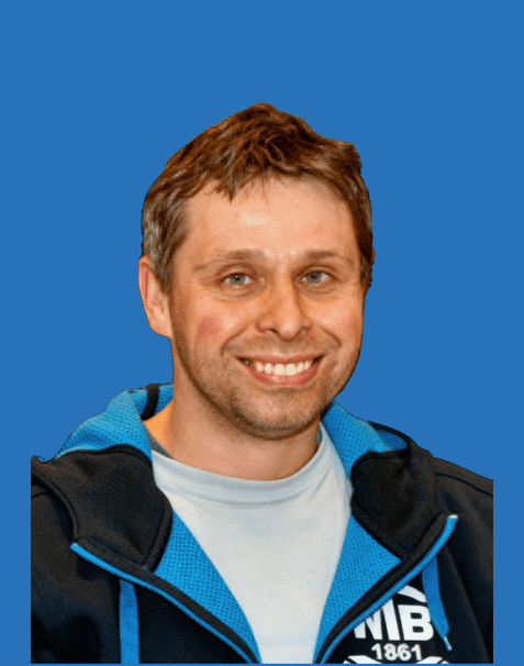Lächelnder Mann mit kurzen, blonden Haaren trägt einen blauen Hoodie vor einem einfarbigen blauen Hintergrund.