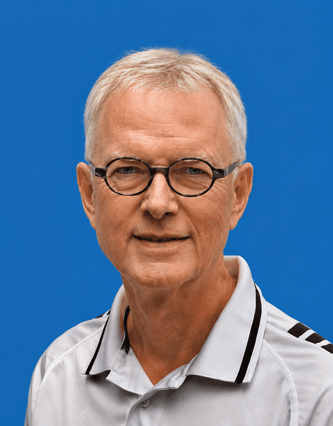 Porträt eines älteren Mannes mit Brille, kurzen grauen Haaren und einem Sporthemd vor einem blauen Hintergrund.