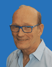 Ein älterer Mann mit blauen Brillengestell und kurzem, grauem Haar lächelt vor einem blauen Hintergrund.