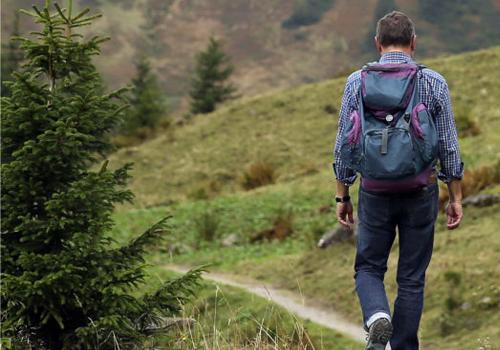 Mann mit Rucksack wandert auf einem schmalen Weg durch eine grüne, bewaldete Landschaft.
