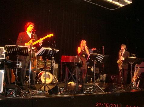 Drei Musiker auf einer Bühne: Sängerin mit Mikrofon, Gitarrist und Saxophonist vor Musik-Equipment.