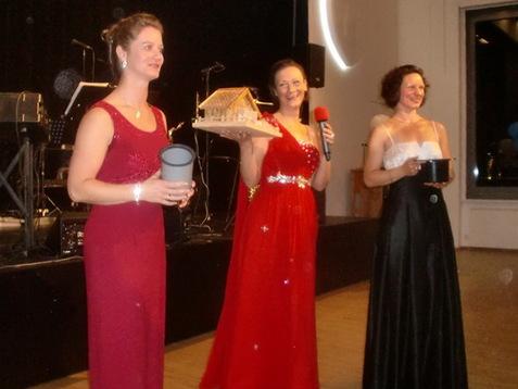 Drei Frauen in eleganten Abendkleidern stehen auf einer Bühne, eine hält ein Hausmodell und eine Mikrofon.