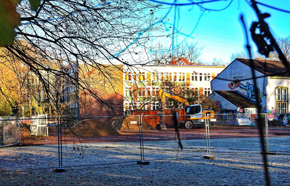 Blick auf einen Baustellenbereich mit Bagger, Unrat und umz&auml;untem Boden vor einem Schulgeb&auml;ude im Hintergrund.