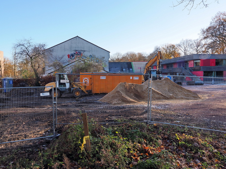 Baugel&auml;nde mit Bagger, Baustellencontainer und Erdhaufen, umgeben von einem Bauzaun und herbstlichen B&auml;umen.