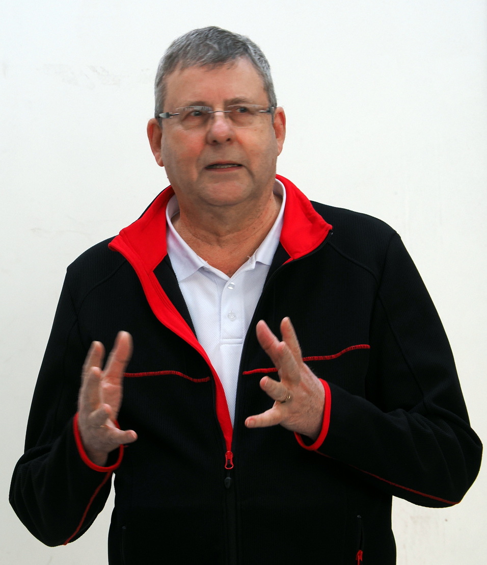 Mann mit grauen Haaren in schwarzer Jacke mit roten Details, spricht und gestikuliert vor wei&szlig;er Wand.