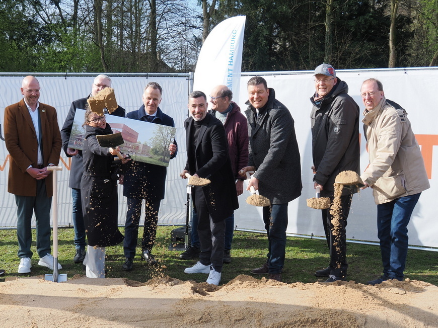 Gruppenbild von mehreren Personen bei der Grundsteinlegung, mit einem Plakat im Vordergrund und Sand, der von Schaufeln f&auml;llt.