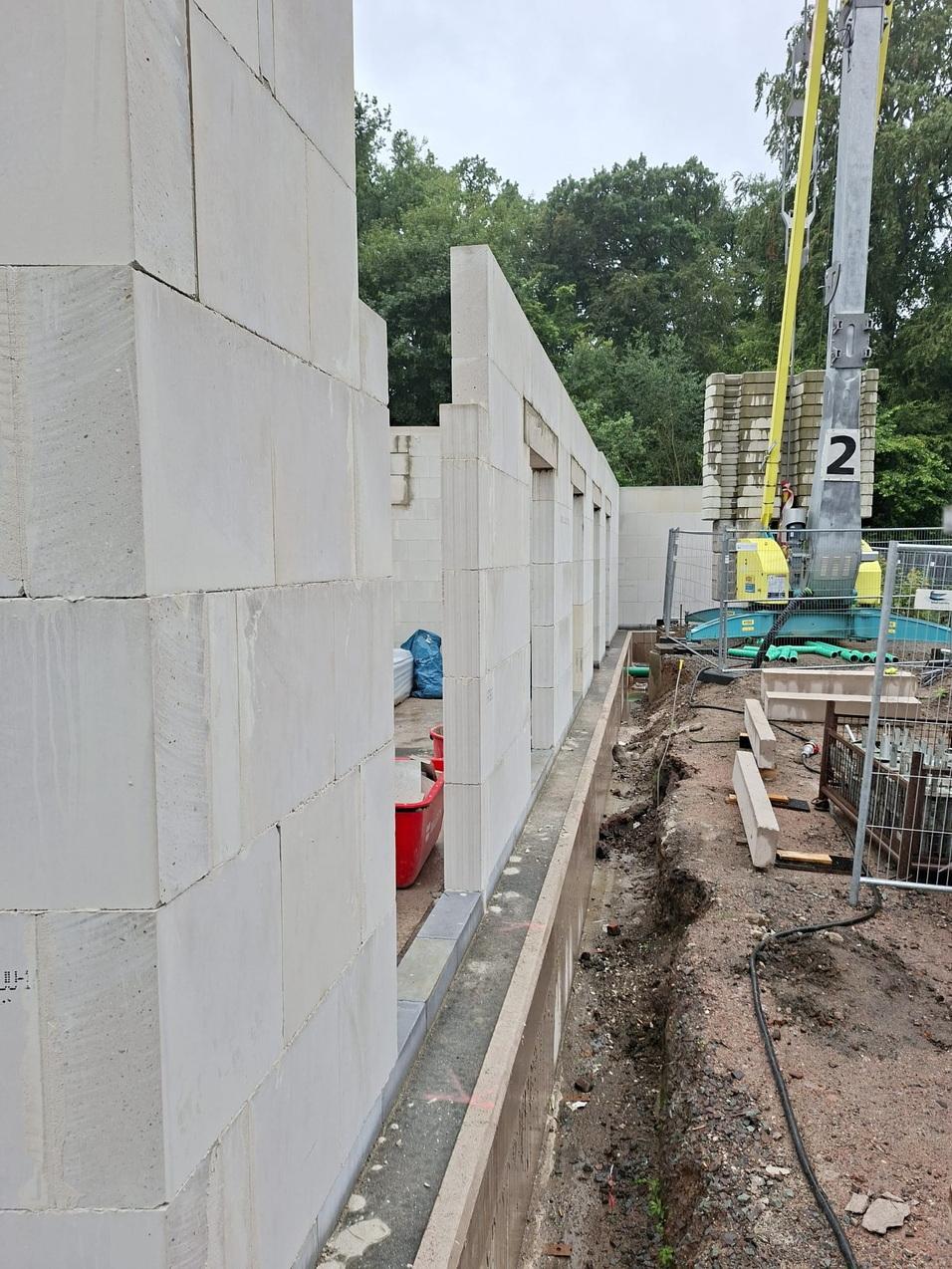 Bau eines Mauerwerks mit weißen Blöcken und offenen Fenstern auf einer Baustelle umgeben von Erde und Baugeräten.