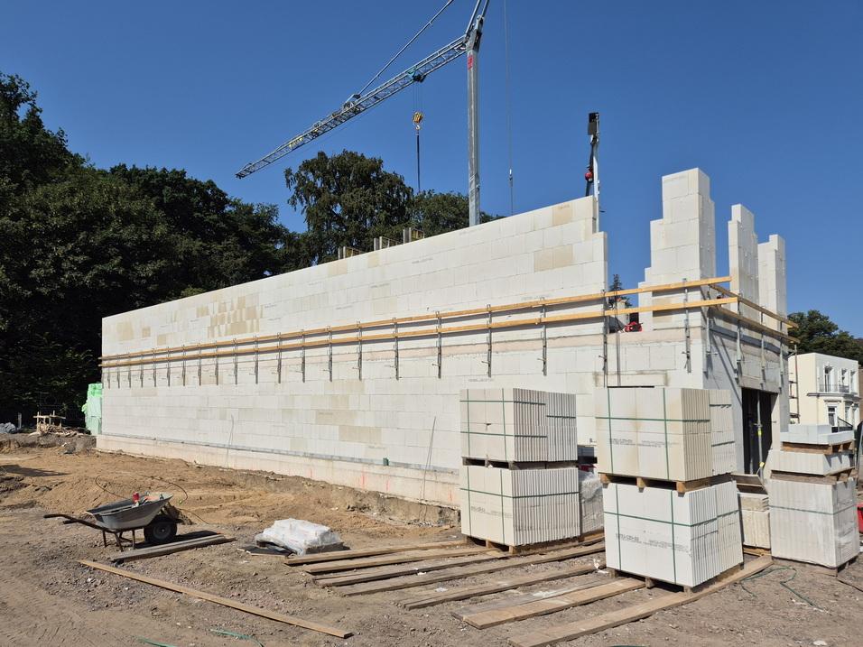 Baustelle mit Mauer aus weißen Steinen, Stapeln von Baumaterial und einem Kran unter blauen Himmel.