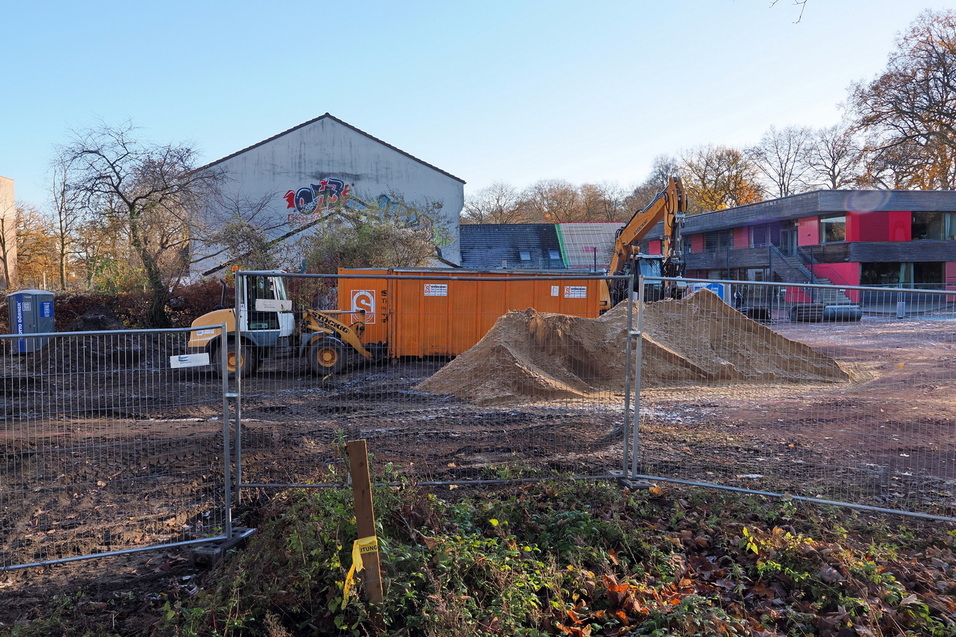 Baugel&auml;nde mit Bagger, Baustellencontainer und Erdhaufen, umgeben von einem Bauzaun und herbstlichen B&auml;umen.