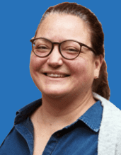 Porträt einer lächelnden Frau mit Brille auf blauem Hintergrund, in blauer Bluse und grauem Cardigan.