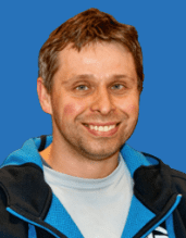 Porträt eines lächelnden Mannes mit kurzen, braunen Haaren vor blauem Hintergrund, bekleidet mit grauem T-Shirt und blauer Jacke.
