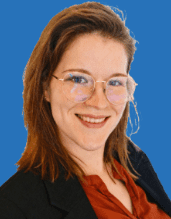 Porträt einer lächelnden Frau mit Brille, die eine schwarze Blazerjacke und ein rotes Oberteil trägt, vor blauem Hintergrund.