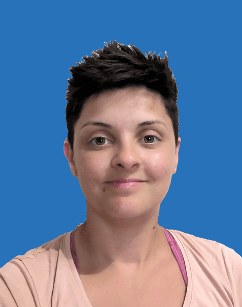 Porträt einer Frau mit kurzem, dunklem Haar und einem Lächeln, vor einem blauen Hintergrund. Sie trägt ein rosa Shirt.