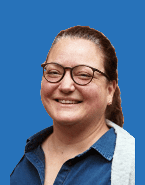 Lächelnde Frau mit Brille und langen braunen Haaren vor blauem Hintergrund, trägt ein Jeanshemd mit grauer Jacke.