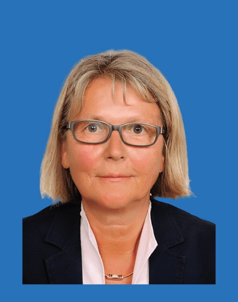 Porträt einer Frau mit blonden, schulterlangen Haaren, Brille und blauem Hintergrund, in Blazer und Bluse gekleidet.