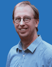 Porträt eines lächelnden Mannes mit Brille, der ein kariertes Hemd trägt, vor einem blauen Hintergrund.