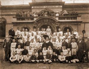 Gruppenfoto von Kindern und Erwachsenen vor einem historischen Gebäude, alle in hellen Uniformen gekleidet.