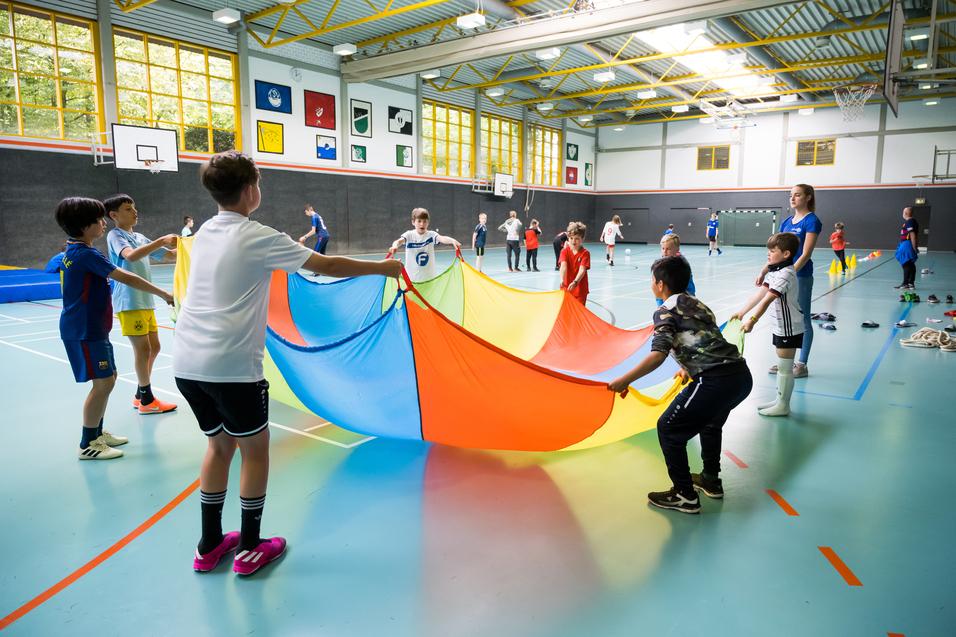 Gruppe von Kindern in einer Sporthalle, die einen bunten Parachute in die Höhe halten und gemeinsam spielen.