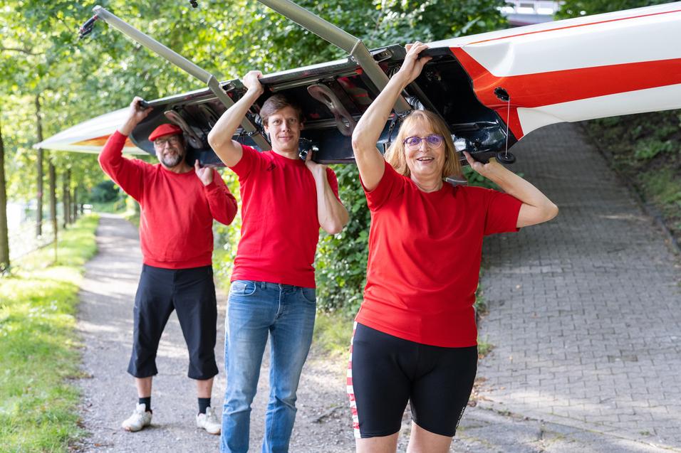 Drei Personen tragen ein Ruderboot auf ihren Schultern, während sie einen Weg entlanglaufen, umgeben von Bäumen.