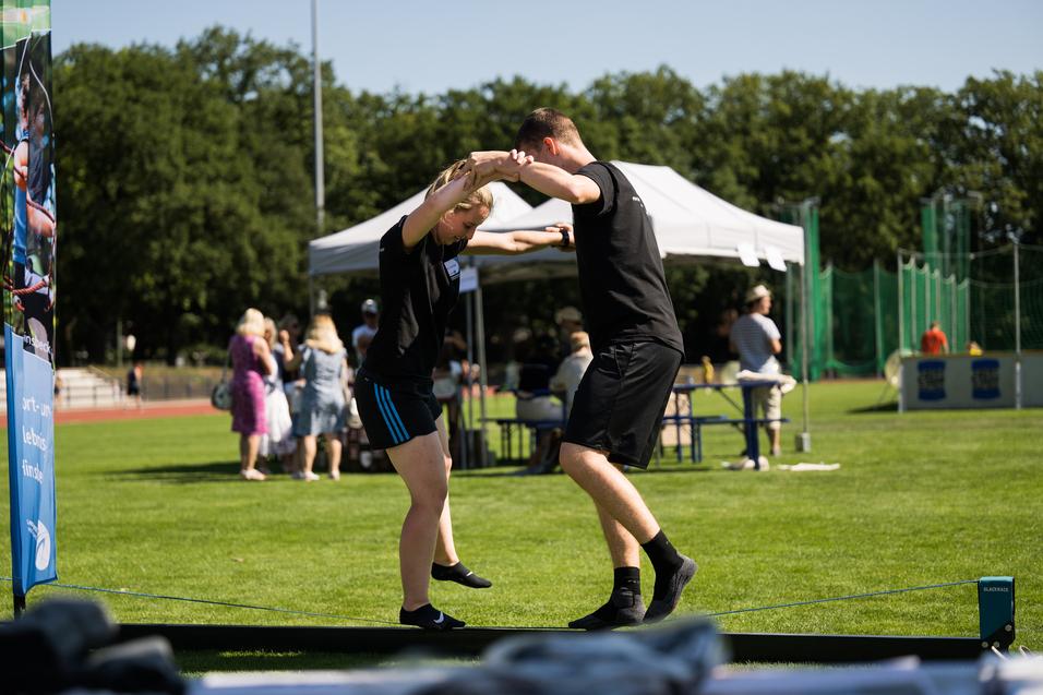 Zwei Personen balancieren auf einem Slackline, umgeben von Zuschauern auf einem Sportplatz.