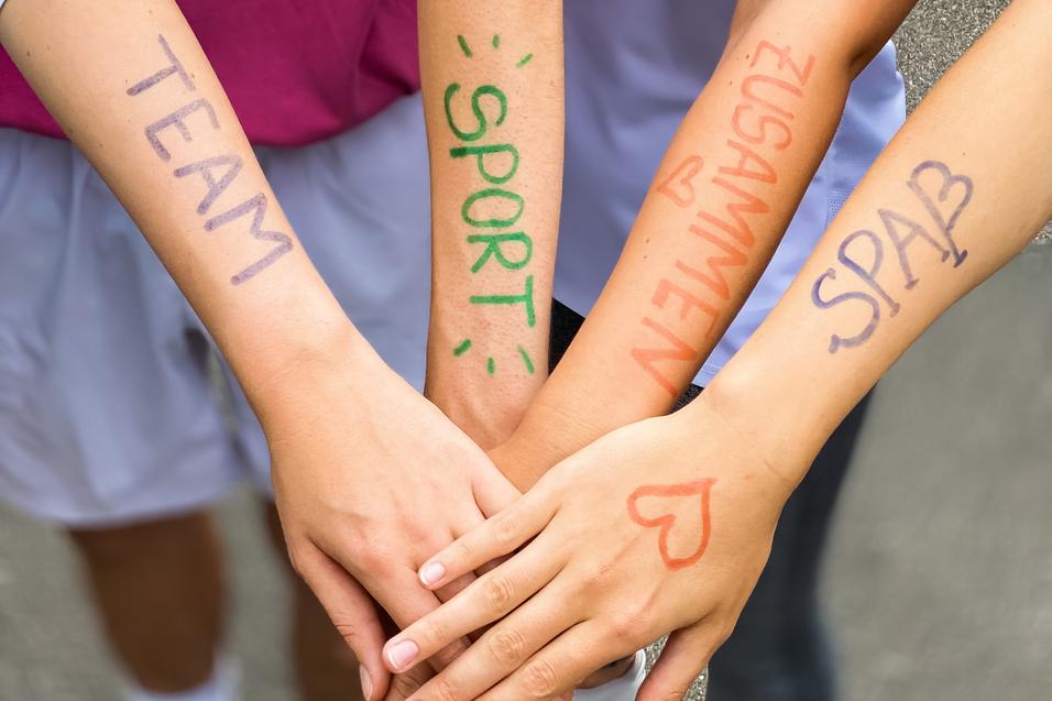 Vier Hände mit bunten Schriftzügen "TEAM", "SPORT", "ZUSAMMEN" und "SPASS" liegen übereinander auf einem grauen Untergrund.