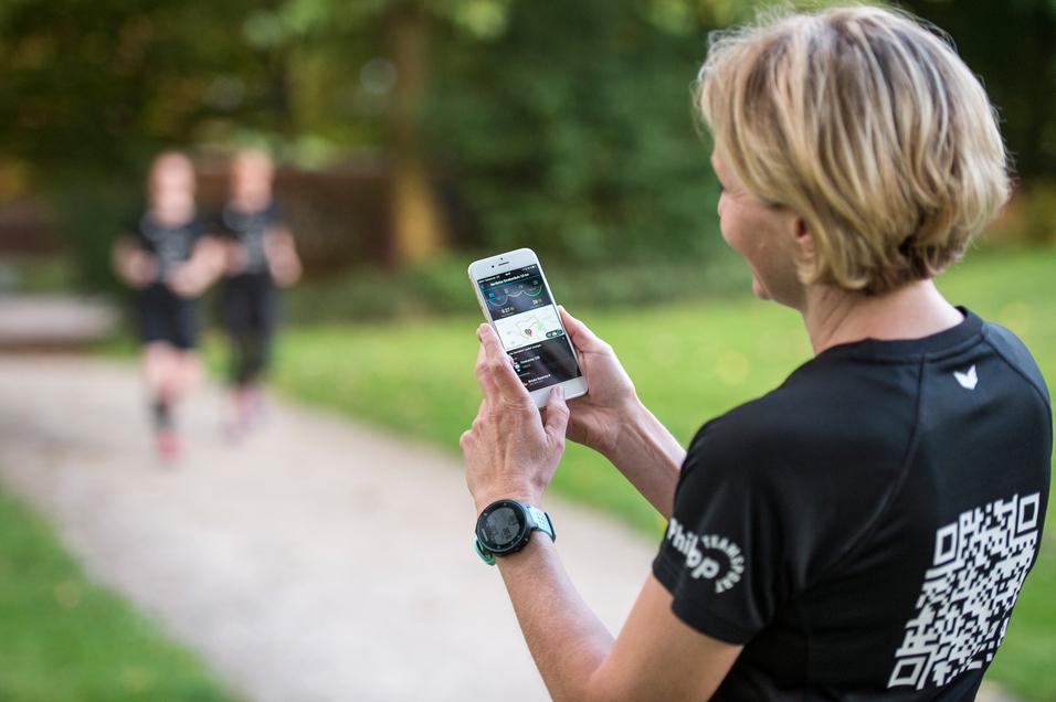 Frau mit kurzem Haar schaut auf ihr Smartphone, während zwei Jogger im Hintergrund auf einem Weg vorbeilaufen.