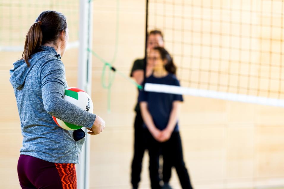 Eine Person mit einem Volleyball steht vor dem Netz, während zwei weitere Personen im Hintergrund warten.